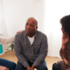 A group of people discussing mental health. The man in the centre has a serious expression while the others look supportive.